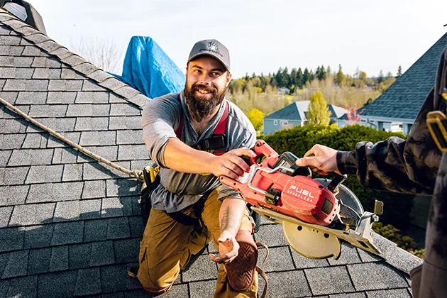 Roofing repair specialist on roof, attached with safety harness, being handed a tool by a co-worker